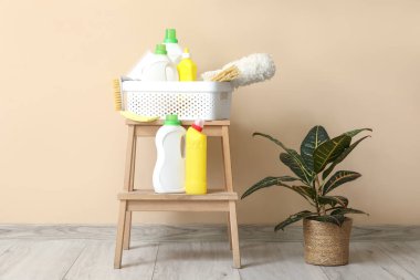Bej duvarın yanındaki merdiven üzerinde temizlik malzemeleri olan plastik sepet.