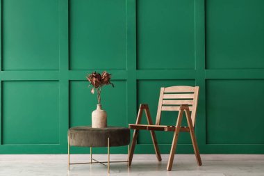 Yeşil duvarın yanındaki vazoda protea çiçekleri olan sandalye ve puf.