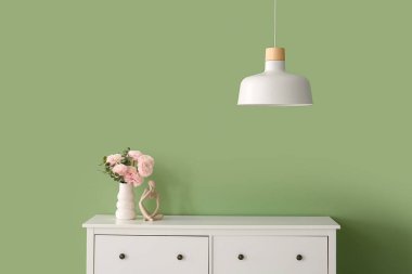 Çekmece dolabında güzel çiçekler ve modern odada yeşil duvarın yanında asılı bir lamba olan bir vazo.