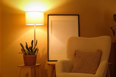 Gece oturma odasının içinde koltuk, merdiven, saksı ve sarı duvarın yanında parlayan lamba var.