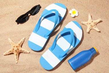 Plaj ayakkabılarıyla kompozisyon, güneş kremi şişesi ve kumun üzerinde güneş gözlüğü, yakın plan.