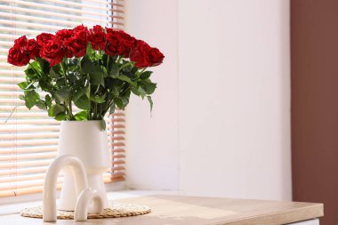 Güzel kırmızı güller ve dekorlu vazo şık bir odanın penceresinin yanındaki masada, yakın plan.