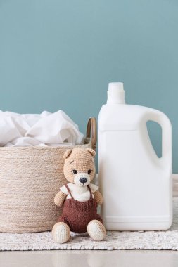 Yeşil duvarın yanındaki halının üzerinde oyuncak ayı ve deterjan olan çamaşır sepeti.