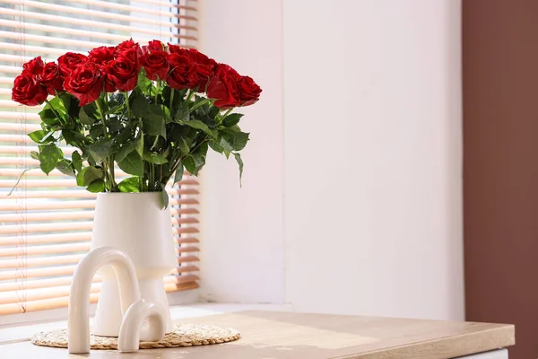 Güzel kırmızı güller ve dekorlu vazo şık bir odanın penceresinin yanındaki masada, yakın plan.