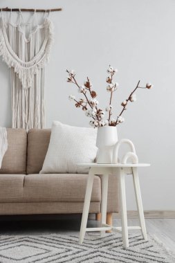 Oturma odasının iç tarafındaki beyaz duvarın yanında bej renkli kanepe, masa ve vazo.