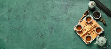 Tepsi, çaydanlık, bir fincan sıcak içecek, kuru ütülenmiş puer yaprakları, bambu hasırı ve maşayla yeşil arka planda yazı için yer, üst görünüm