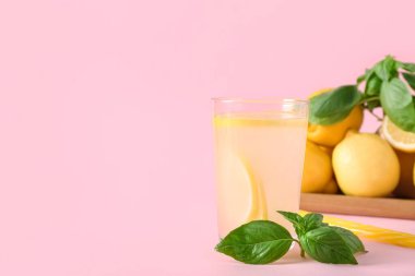 Bir bardak taze limonata, fesleğen yaprakları ve pembe arka planda limonlar.