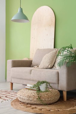 Ev bitkileriyle süslü hasır puf ve oturma odasındaki yeşil duvarın yanındaki rahat kanepe.