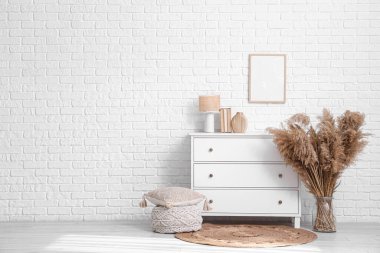 Yumuşak yastıklı şık örülmüş bir pouf ve beyaz tuğla duvarın yanındaki çekmeceler.
