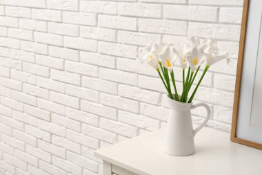 Vase with artificial lily flowers on shelf near white brick wall