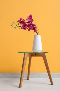 Vase with artificial orchid flower on table near orange wall