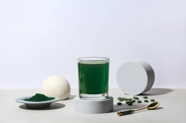 Glass of spirulina drink with pills, spoon, powder and plaster podiums on light background