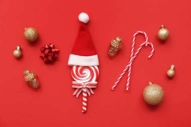 Composition with golden Christmas balls, lollipop and Santa hat on red background