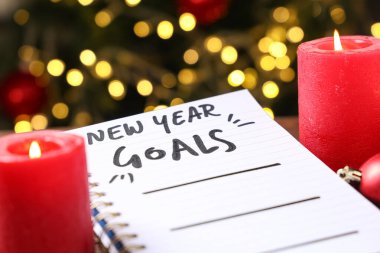 Notebook with New Year goals and burning candles against blurred lights, closeup
