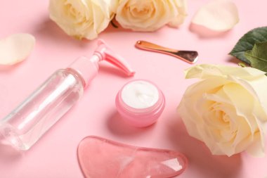 Beautiful white rose flowers with cosmetic products and guasha tool on pink background, closeup