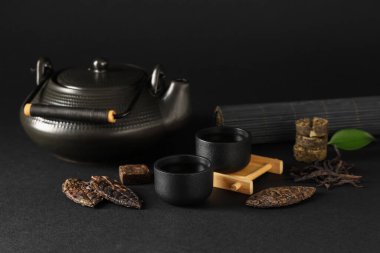 Siyah zemin üzerinde kuru ütülenmiş puer çayı, fincanlar ve çaydanlık bulunan bambu hasırı.