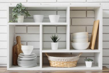 Mutfakta beyaz tuğla duvarın yanında bulaşık ve yapay bitkiler bulunan raf ünitesi.