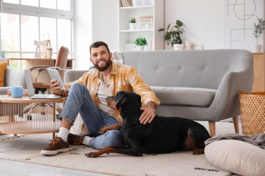 Evde tabletli genç adam ve Dobermann köpeği var.