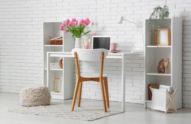 Modern dizüstü bilgisayarlı şık çalışma alanı, bir buket güzel lale çiçeği ve beyaz tuğla duvarın yanındaki raf üniteleri.