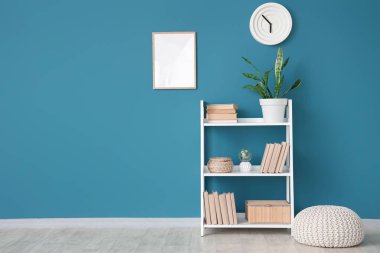 Kitaplı raf ünitesi, ev bitkisi ve mavi duvarın yanında puf.