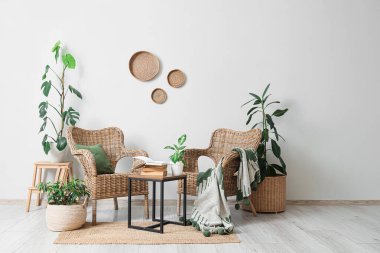 Hasır koltuklar, ev bitkileri, merdivenler ve beyaz duvarın yanındaki sehpanın üzerindeki kitaplarla dolu bir oda.