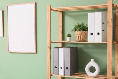 Ofiste yeşil duvarın yanında klasörleri ve yapay bitkileri olan raf ünitesi.