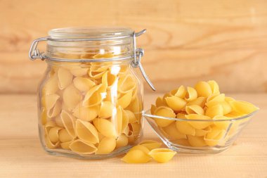 Kavanoz ve cam kase, ahşap arka planda çiğ koni makarnası.