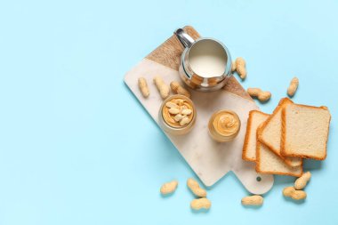 Fıstık ezmeli kavanozlar, bir sürahi süt, kızarmış ekmek ve fındıklar mavi arka planda.