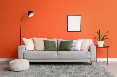 Comfortable grey sofa with soft pillows and floor lamp near orange wall in living room