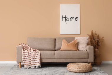 Oturma odasındaki bej duvarın yanında hasır pouf ve pampa otlu rahat kahverengi kanepe.