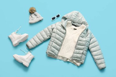 Çizmeli, kışlık şapkalı, süveterli ve mavi arka planda güneş gözlüklü şık kısa gri kabarık ceket.