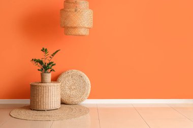 Pouf, ev bitkisi ve hasır masa turuncu duvarın yanında oturma odasının içinde.