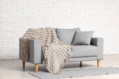 Comfortable grey sofa with plaid near white brick wall in interior of living room