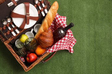 Rattan picnic basket with tableware, bottle of wine, croissant, baguette and fruits on green background