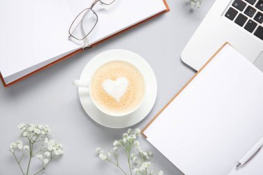 Cup of coffee with eyeglasses, notebooks, laptop and gypsophila flowers on grey background