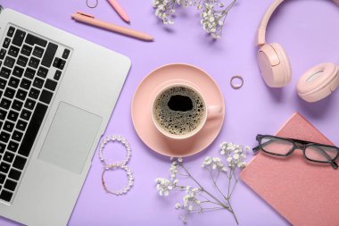 Dizüstü bilgisayarlı, defterli, kulaklıklı, gözlüklü, mücevherli ve mor arkaplanlı bir fincan kahve.