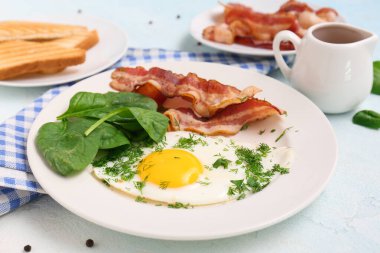Plate with tasty fried egg, spinach, dill and bacon on light blue background