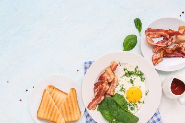Plates with tasty fried egg, spinach, dill, bacon, toasts and jug of ketchup on light blue background