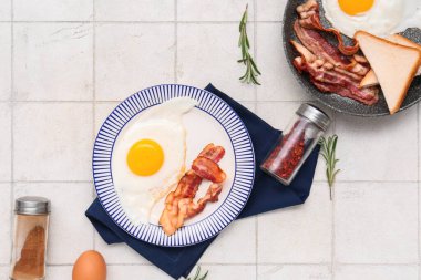 Plate and frying pan with tasty eggs, bacon, toasts, rosemary and spices on white tile background