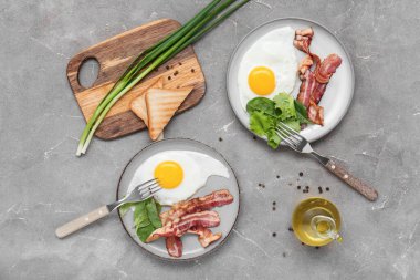 Plates with tasty fried eggs, bacon, salad mix, toasts, green onion and jug of oil on grey grunge background