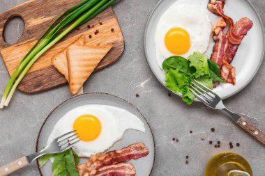 Plates with tasty fried eggs, bacon, salad mix, toasts, green onion and jug of oil on grey grunge background