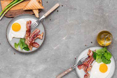 Plates with tasty fried eggs, bacon, salad mix, toasts, green onion and jug of oil on grey grunge background