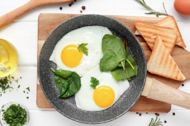 Frying pan with tasty eggs, spinach, dill, rosemary, toasts and jug of oil on white wooden background