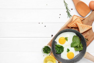 Frying pan with tasty eggs, spinach, dill, rosemary, toasts and jug of oil on white wooden background