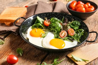 Frying pan with tasty fried eggs, tomatoes, salad mix and toasts on wooden background