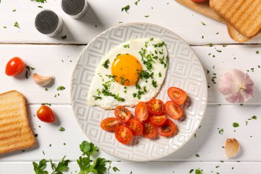Plate with tasty fried egg, parsley, tomatoes, garlic and toasts on white wooden background
