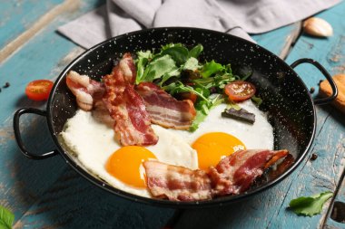 Frying pan with tasty fried eggs, salad mix, bacon and tomatoes on wooden background