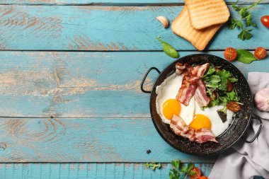 Frying pan with tasty fried eggs, salad mix, bacon, tomatoes, garlic and toasts on wooden background