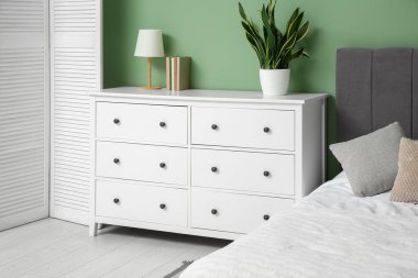 White chest of drawers with houseplant and lamp near bed in bedroom