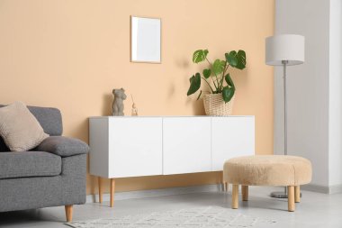 Oturma odasındaki şık kanepenin yanında çiçekli beyaz çekmece ve puf.